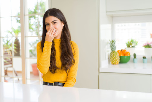 A woman sitting in a kitchen and holding her nose because of a smelly air conditioner.