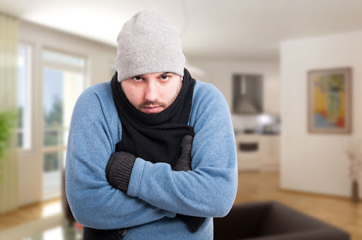 The image shows a man wearing a blue sweater and a red scarf in a cold house without effective heating.
